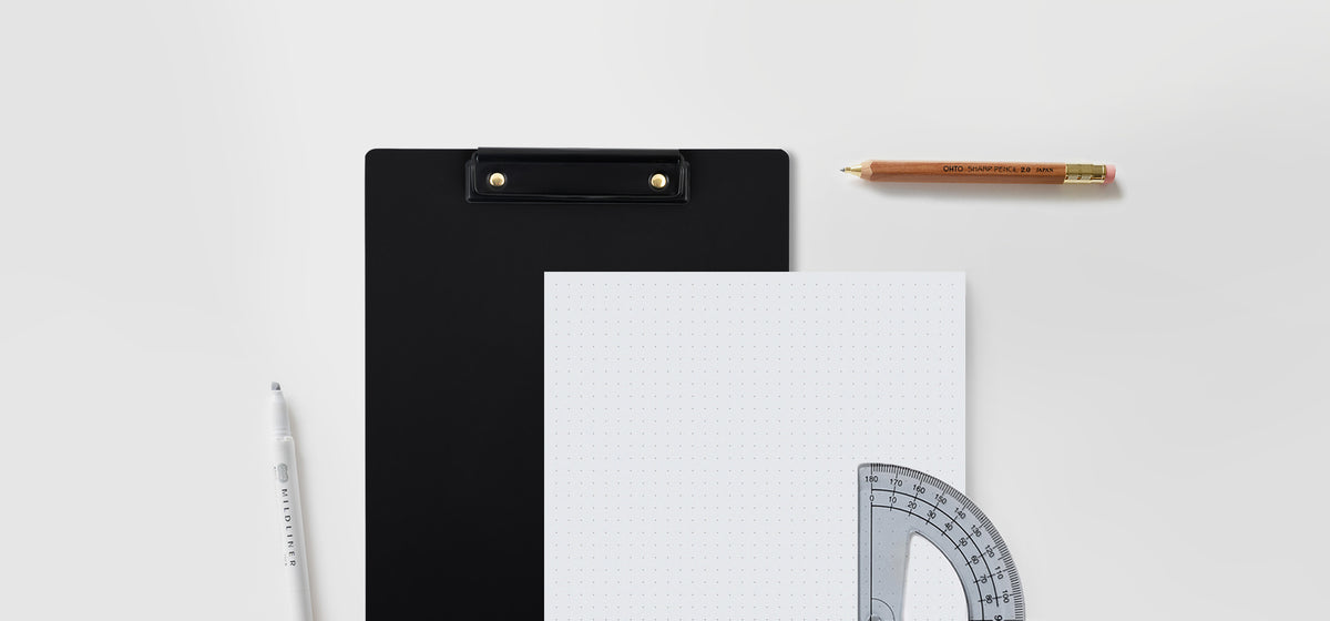 The Appointed Dot Grid Pad and Writing Clipboard, from the brand new Workspace Collection, sit against a gray background with a pencil, protractor, and highlighter aside.