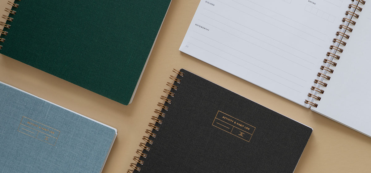 The Activity & Habit Log in a few different colors lies in a grid pattern against a neutral beige background. An entry spread is seen in the upper corner where the log lies open