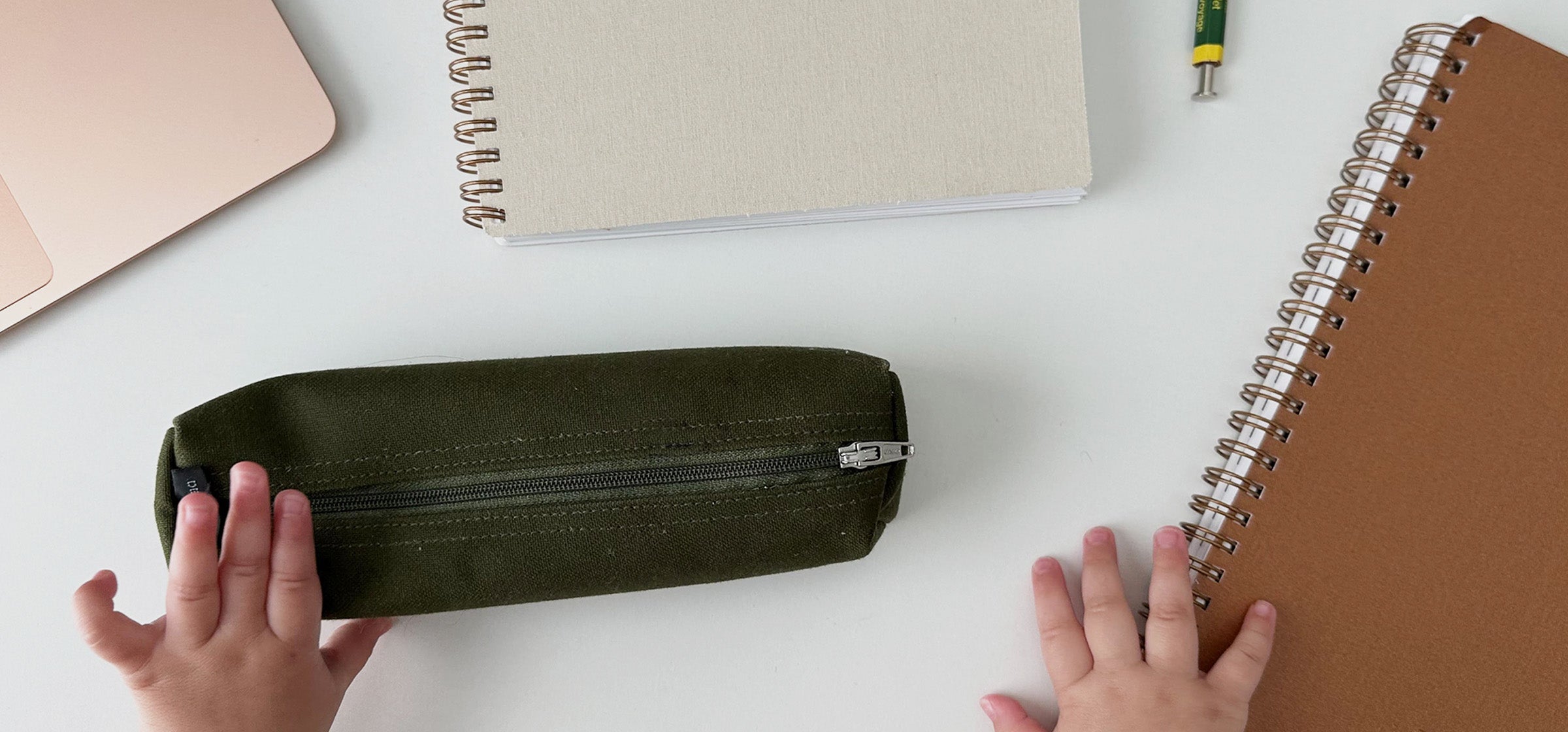 Appointed planners, workbooks, and a pencil case are strewn across a white table top. A baby's hand reaches out to grasp the pen case and planner.