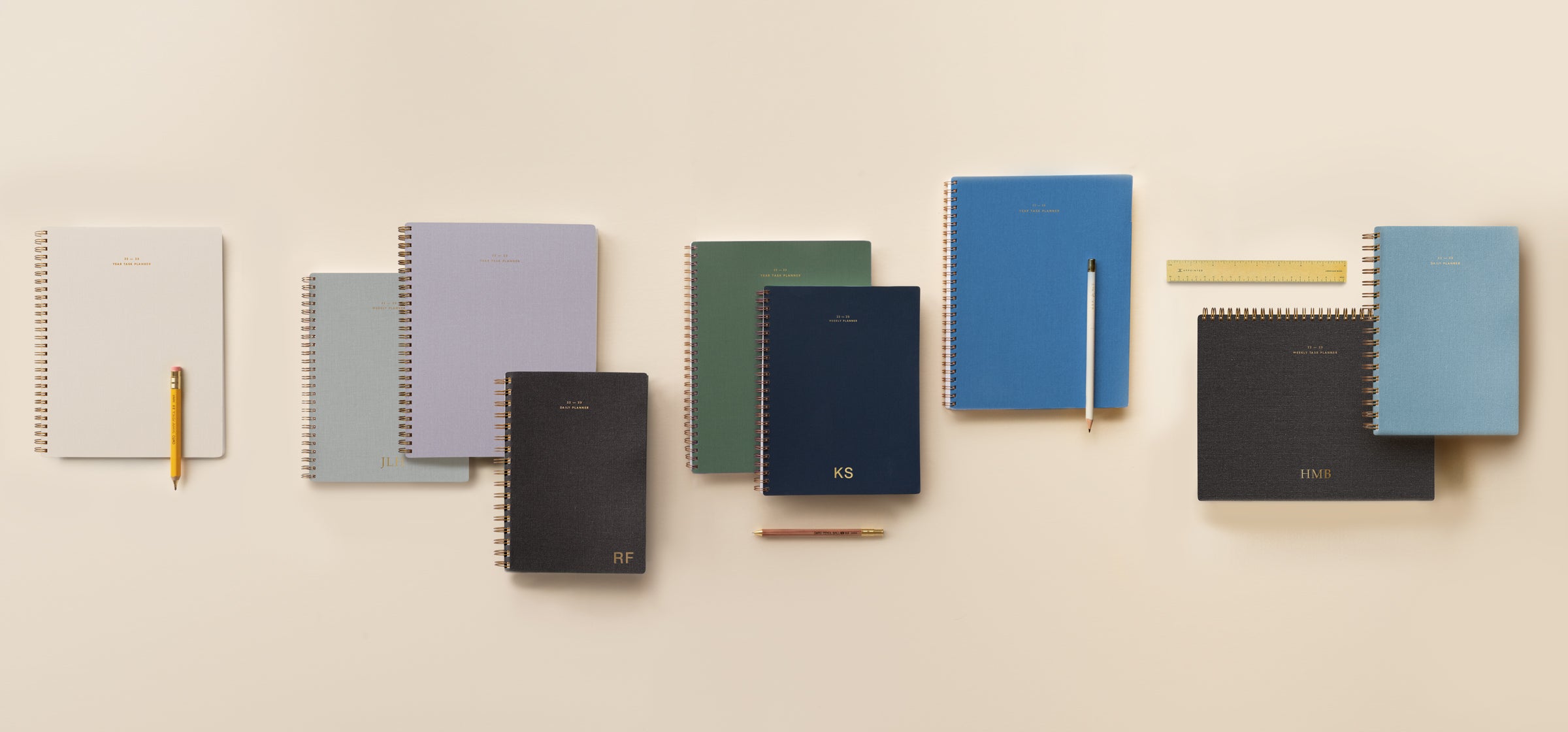 An assorted collection of 22-23 planners lies in an imperfect horizontal row across a light peach background. A few accessories including pencils, pens, and a brass ruler are interspersed.