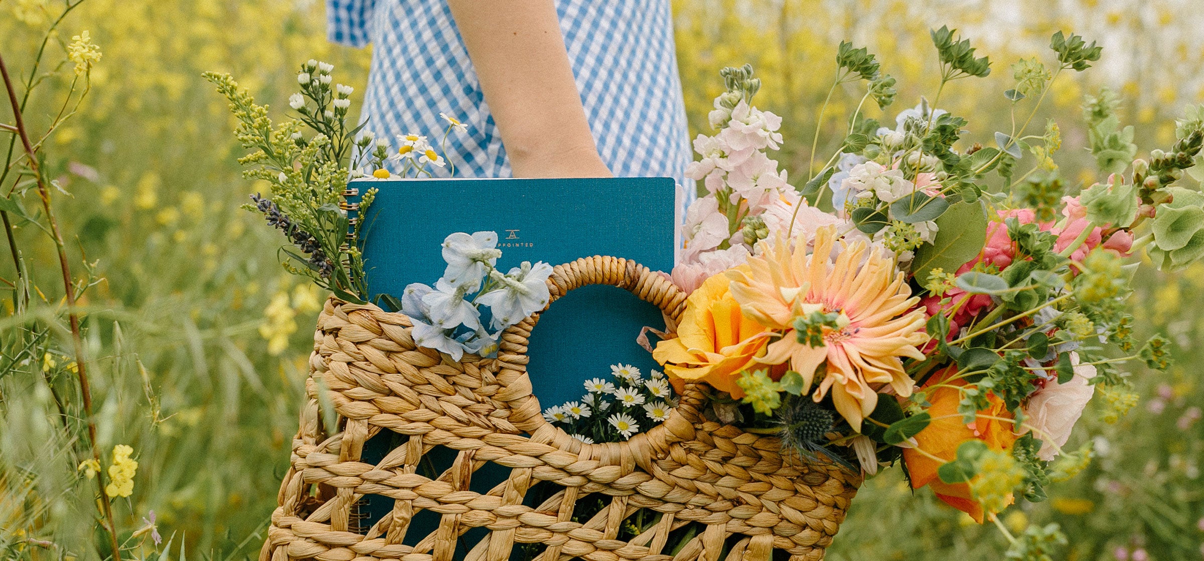 A woman in a blue gingham dress stands in a field of grass and flowers. You can see her from the waist down, as she holds a straw basket with wooden handle filled with wildflowers. The Atlas Blue Appointed Notebook peeks out the top of the basket.