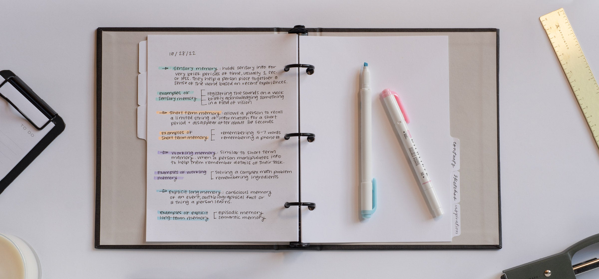 The Compact Binder lies open on a white desktop. Class notes are filled in on the left-hand side of the lined pages. Highlighters sit on top of the planner and other accessories are strewn to the side including a brass ruler and mini clipboard