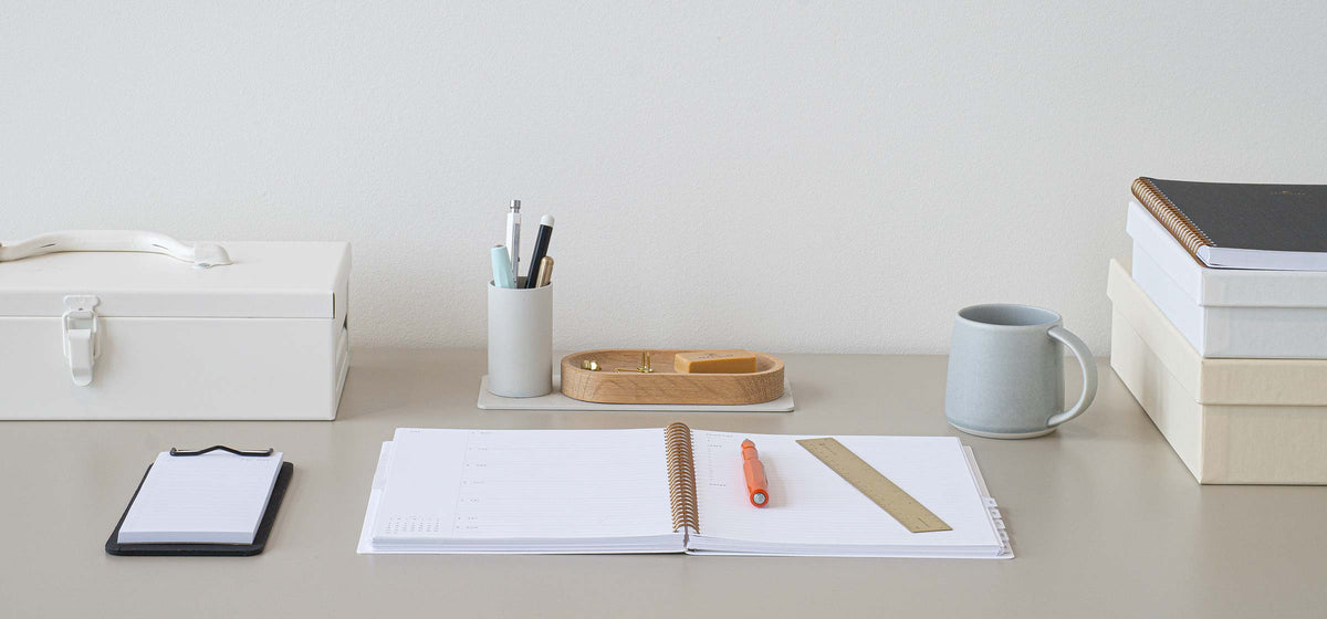 A serene desk images shows an open year task planner on a cool-toned wooden desk. Various accessories also sit on the desk including a steel toolbox, mini clipboard, pen cup, a ceramic mug, and books.