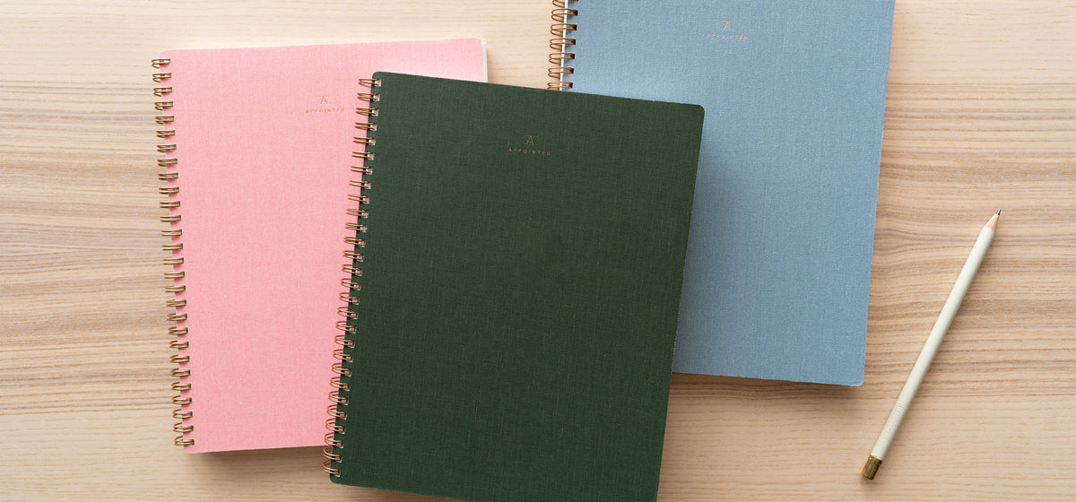 Blossom Pink, Hunter Green, and Chambray Blue Notebooks sit on top of a wood desk with a no. 2 pencil aside them