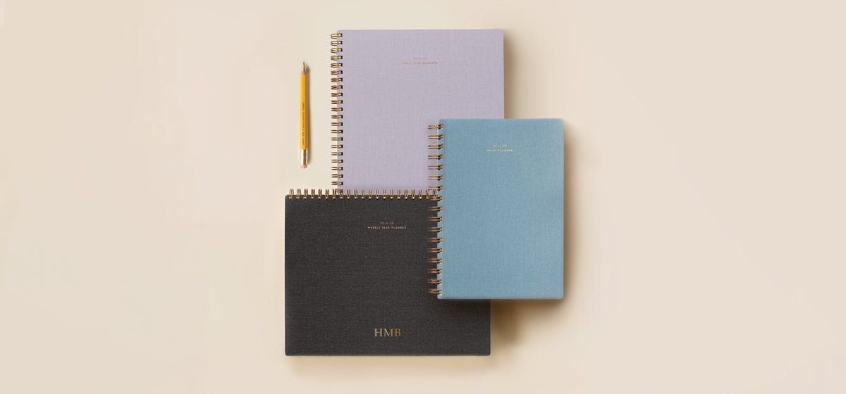 New 22-23 Appointed planners are staggered and stacked against a light pink background including a year task planner in lavender gray, wire daily planner in chambray blue, and weekly task planner in hunter green