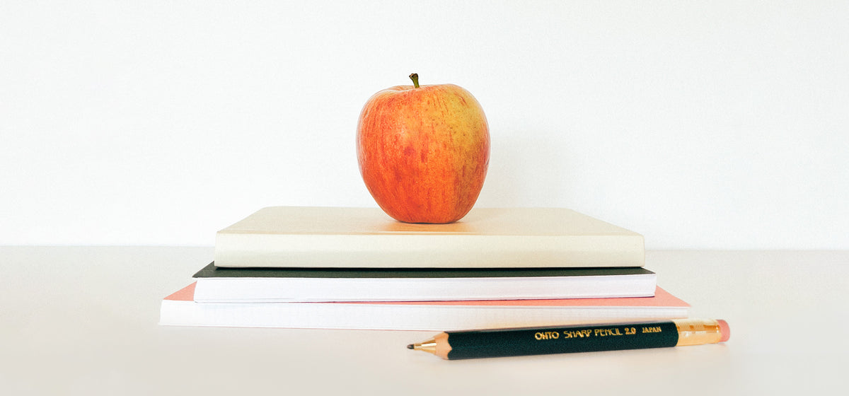 A bright red apple sits on top of a stack of notebooks on a beige table. A thick black wooden mechanical pencil sits in front of the notebook stack.